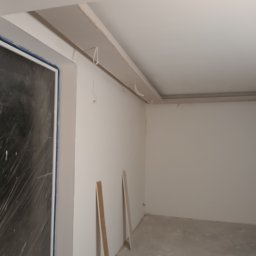 BartexDesign - Wnętrze w trakcie prac wykończeniowych: ściany po szpachlowaniu, sufit z elementami dekoracyjnymi, okno zabezpieczone folią. Dwie listwy przypodłogowe oparte o ścianę.