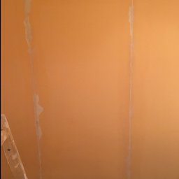 BartexDesign - Pomarańczowa ściana w trakcie szpachlowania, widoczne ślady po taśmie malarskiej i szpachli, fragment drabiny w lewym dolnym rogu.