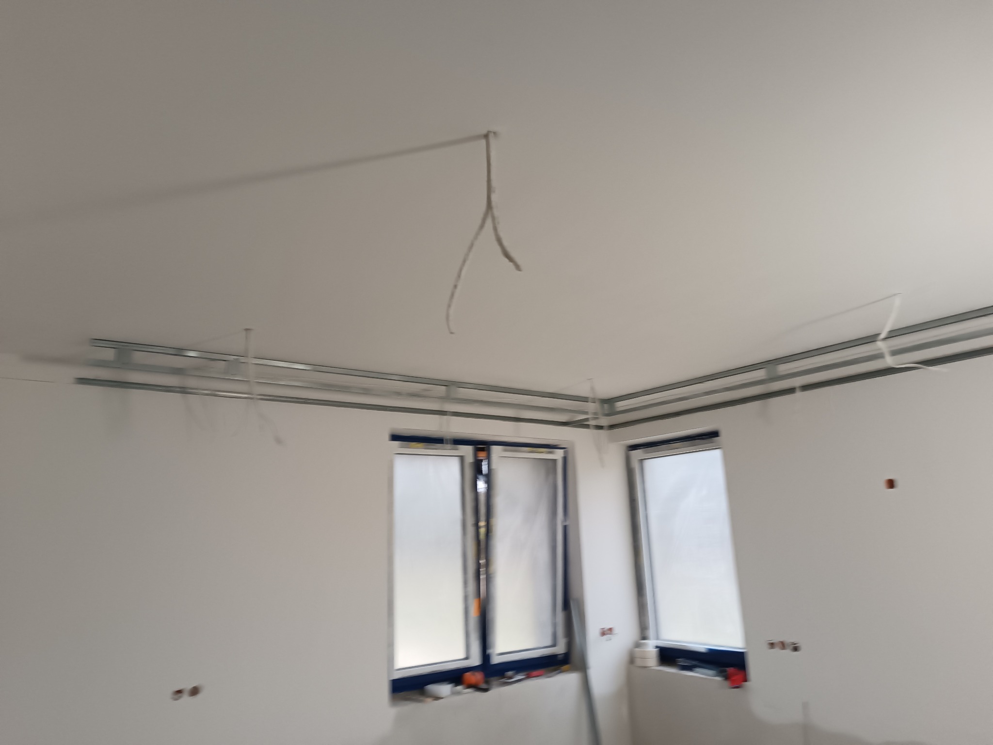 Montaż metalowej konstrukcji sufitu podwieszanego w jasnym pokoju z dwoma oknami. Widoczne profile i przewody elektryczne.