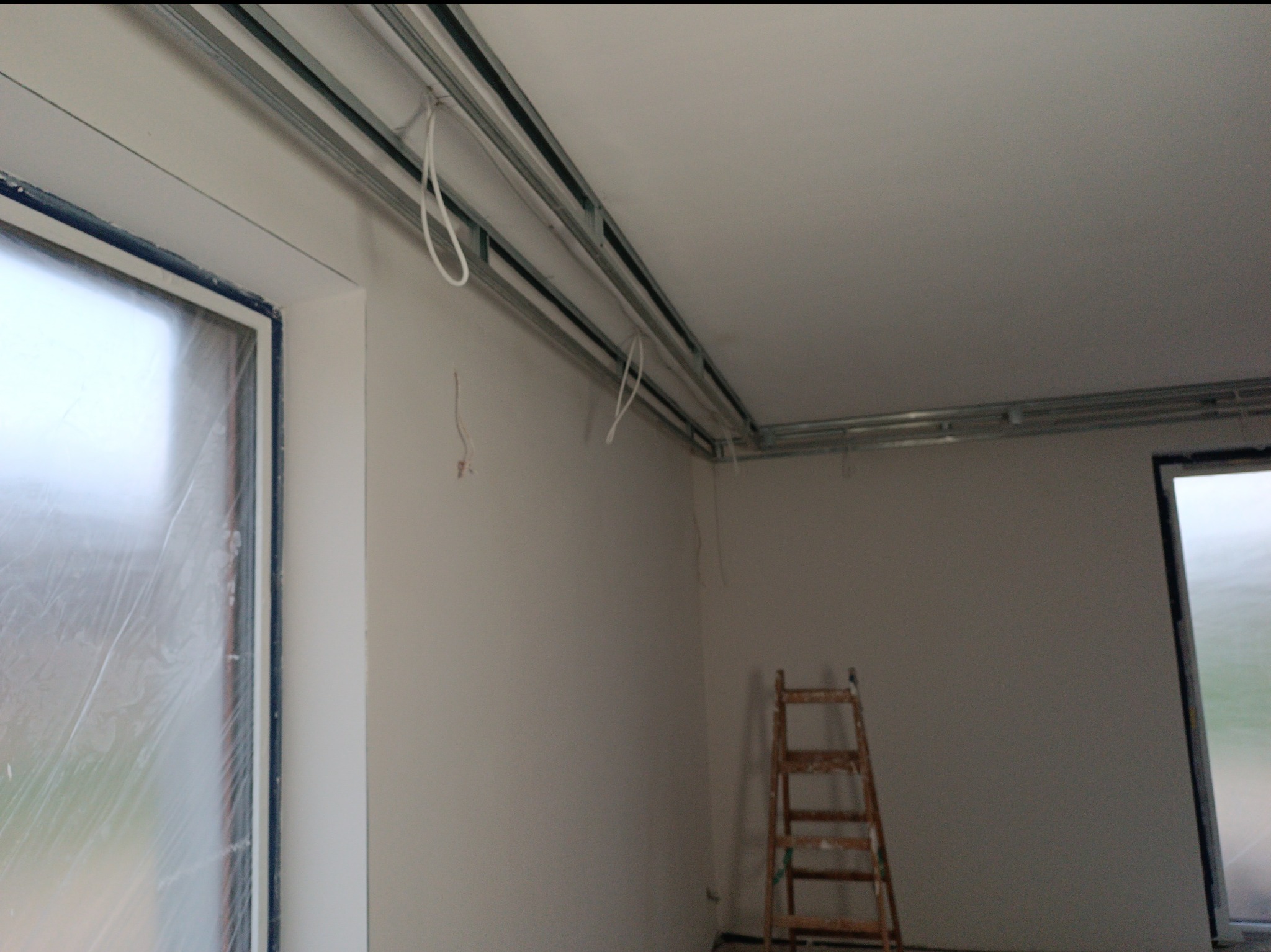 Montaż stelaża sufitu podwieszanego z płyt karton-gips. Widoczne profile, przewody i drabina. Okno zabezpieczone folią. Ściany przygotowane do dalszych prac.