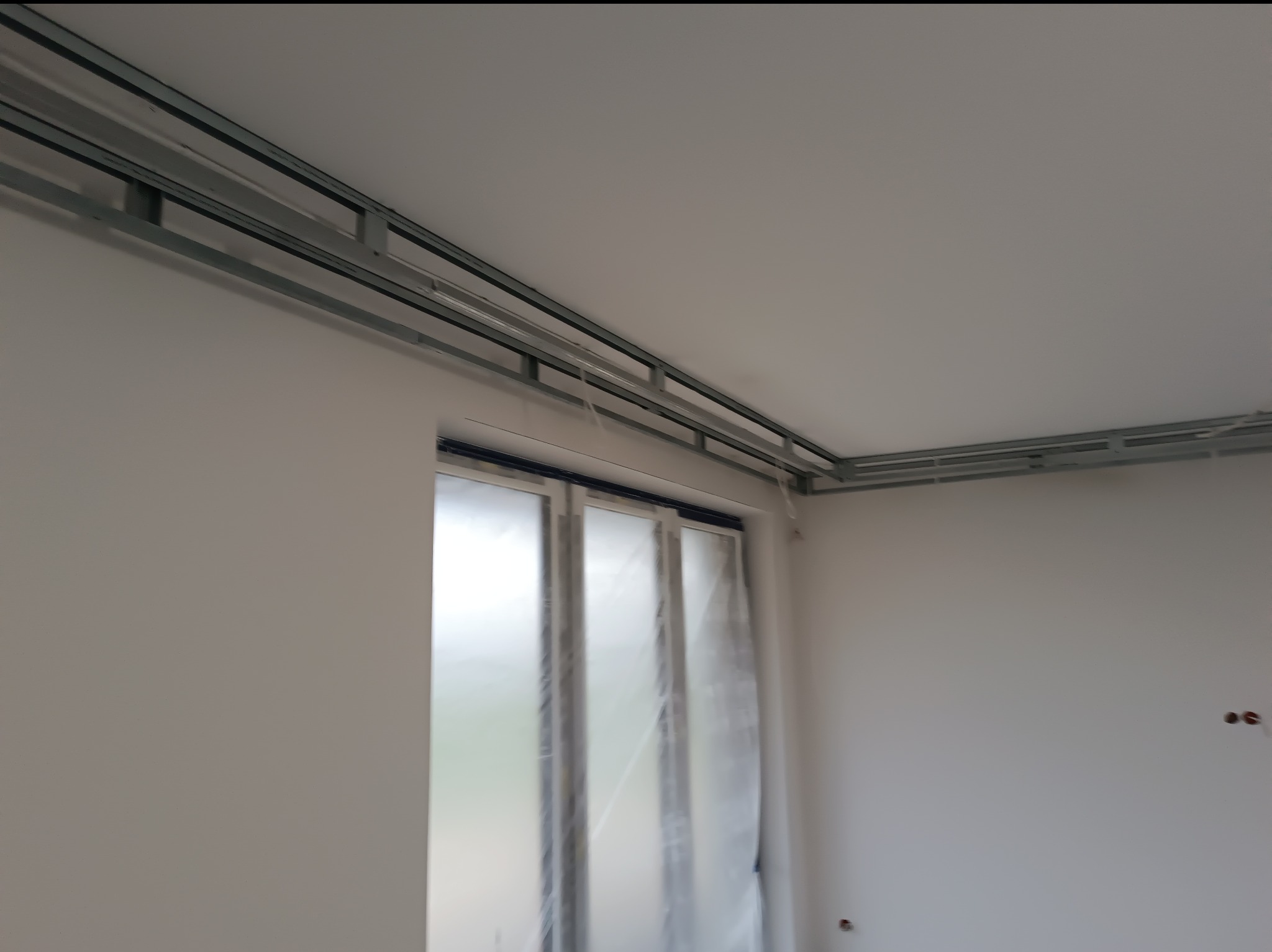 Montaż metalowej konstrukcji sufitu podwieszanego w jasnym wnętrzu. Widoczne profile przy ścianach i oknie zabezpieczonym folią. Etap prac wykończeniowych.