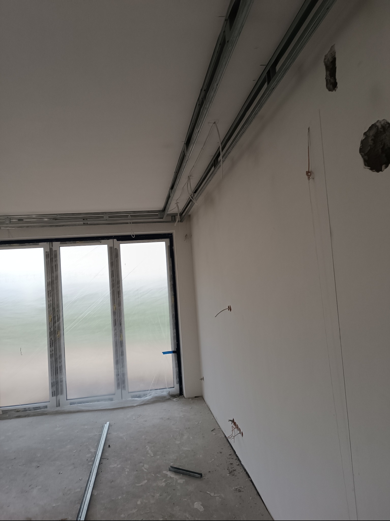 Surowe wnętrze z metalową konstrukcją sufitu podwieszanego i dużym oknem balkonowym. Widoczne przewody elektryczne i niedokończone ściany.