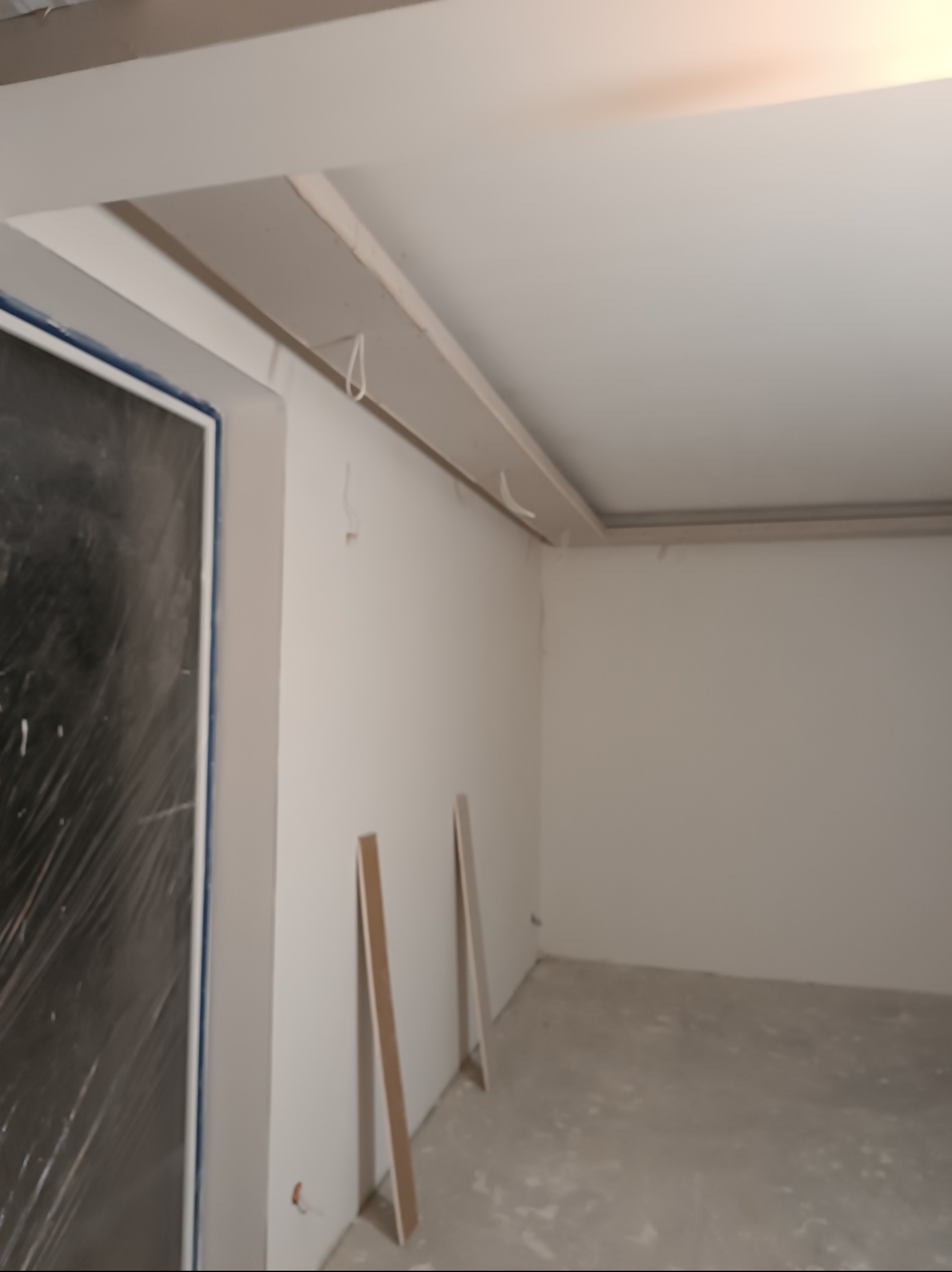 Wnętrze w trakcie prac wykończeniowych: ściany po szpachlowaniu, sufit z elementami dekoracyjnymi, okno zabezpieczone folią. Dwie listwy przypodłogowe oparte o ścianę.