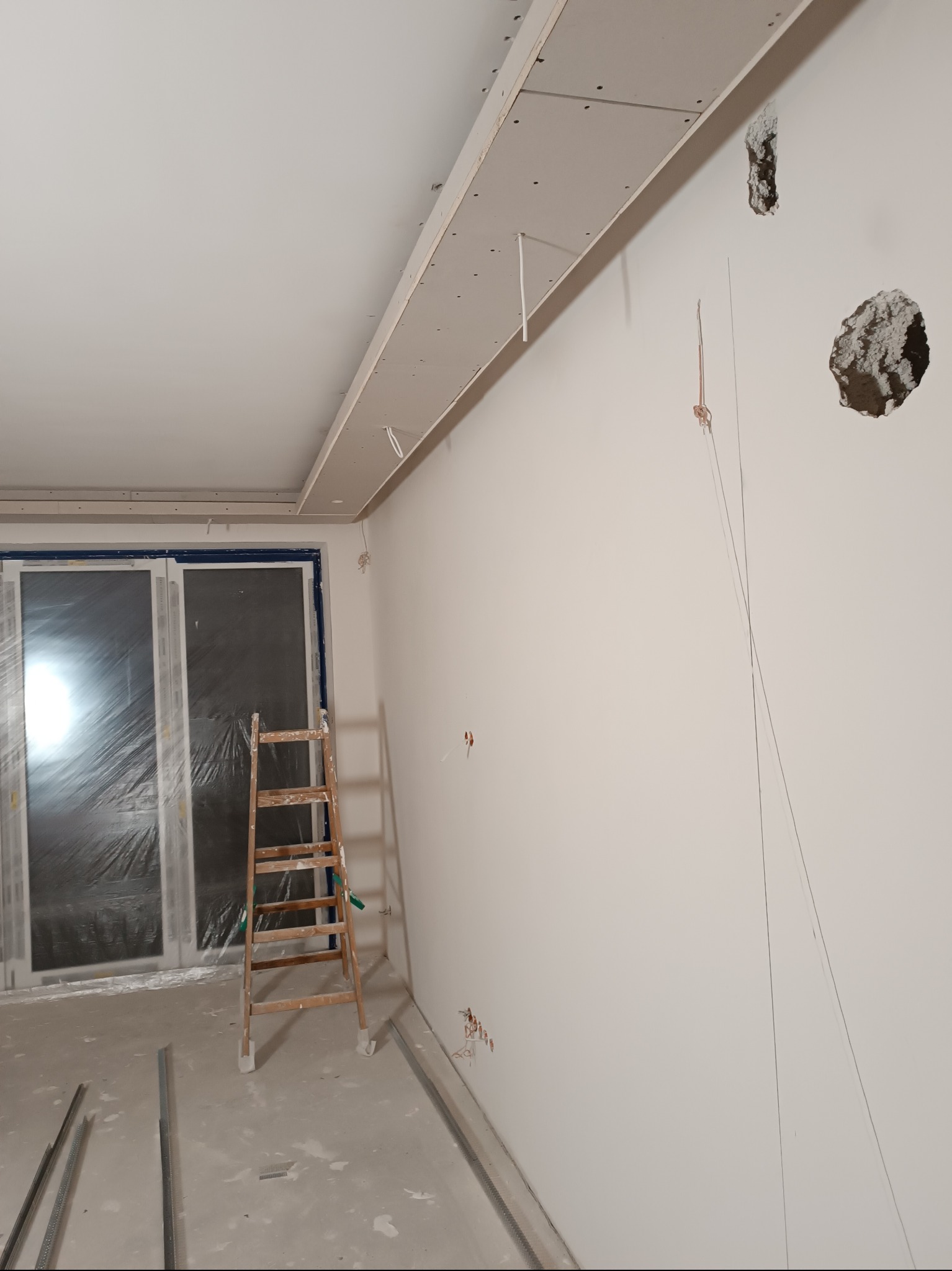 Montaż sufitu podwieszanego z płyty gipsowej w jasnym wnętrzu. Widoczne profile, drabina i przygotowane otwory w ścianie. Okno zabezpieczone folią malarską.