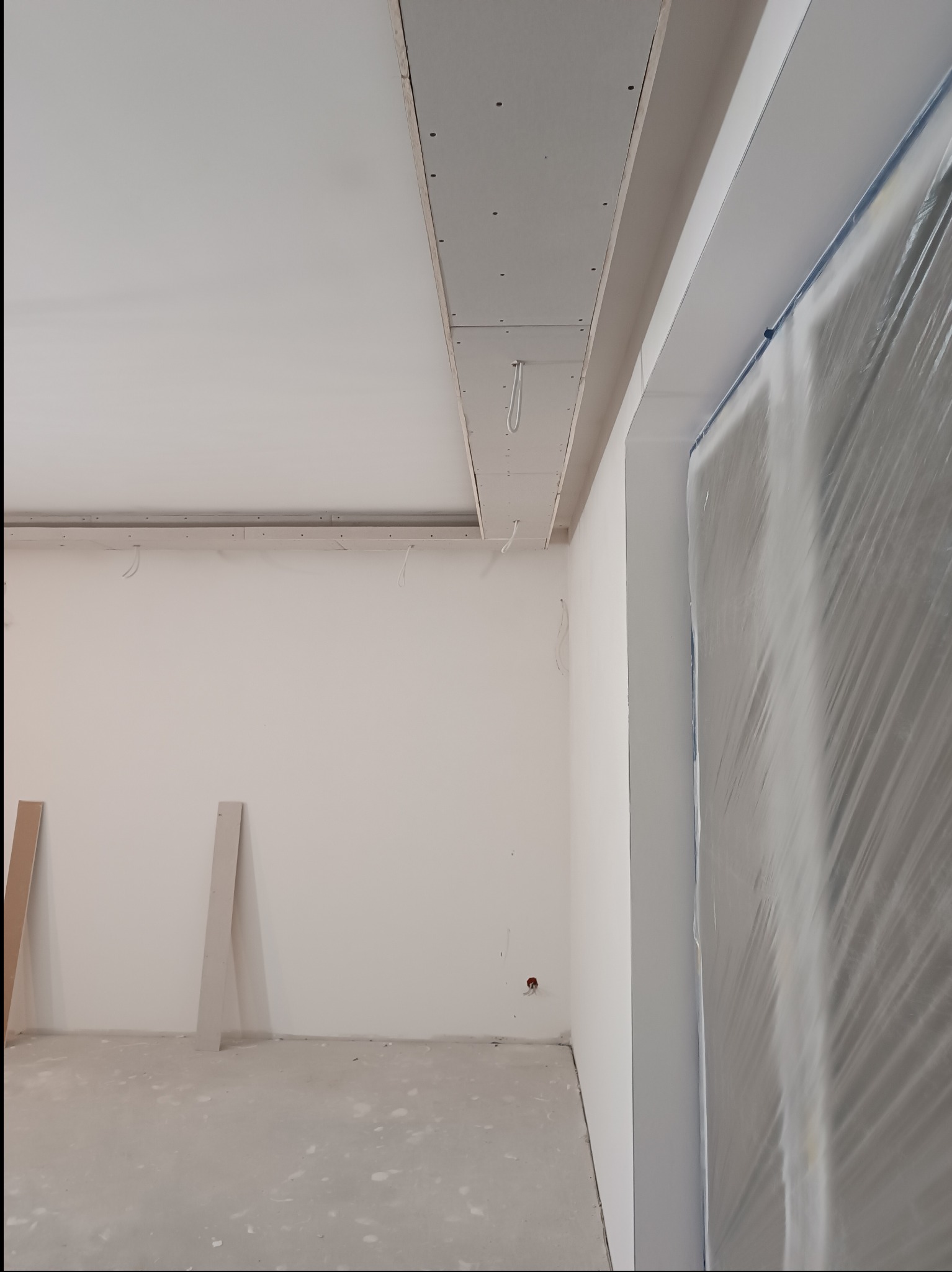 Wnętrze w trakcie prac wykończeniowych: sufit podwieszany z płyt karton-gips, zabezpieczone folią drzwi, ściany przygotowane do malowania, widoczne kable.