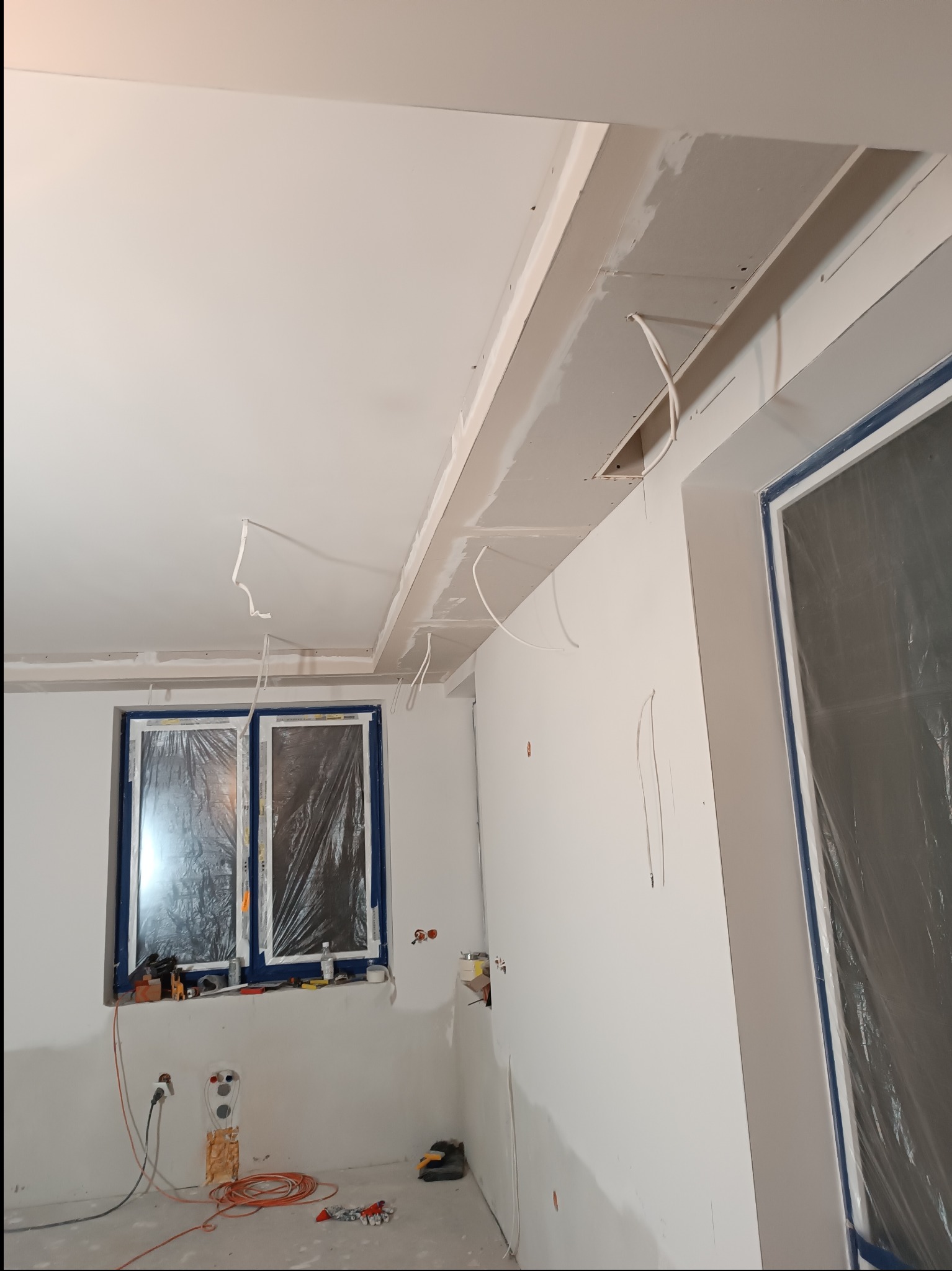 Wnętrze w trakcie prac wykończeniowych: zamontowana płyta karton-gips na suficie, okno zabezpieczone folią, widoczne przewody elektryczne i narzędzia.