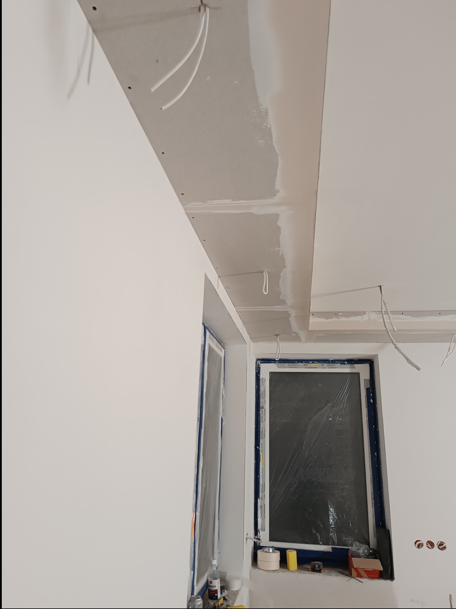 Wnętrze w trakcie prac wykończeniowych: ściany i sufit z płyt karton-gips, częściowo wyszpachlowane, zabezpieczone okno folią malarską. Widoczne kable elektryczne.