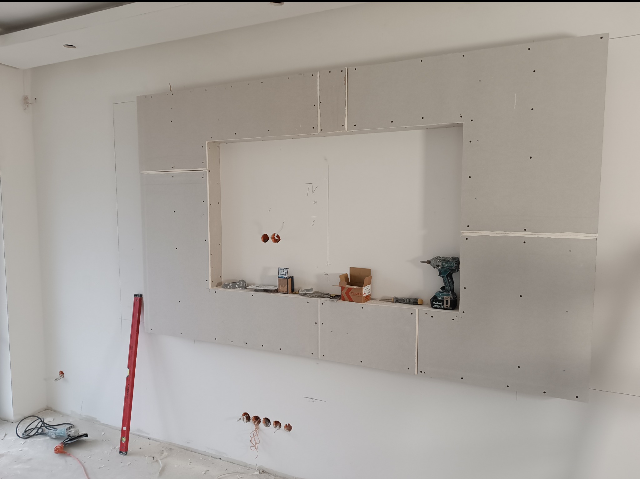 Montaż płyty karton-gips na ścianie, tworzącej ramę pod telewizor. Widoczne narzędzia, poziomica i otwory pod instalacje. Praca w trakcie realizacji, surowy stan wykończenia.