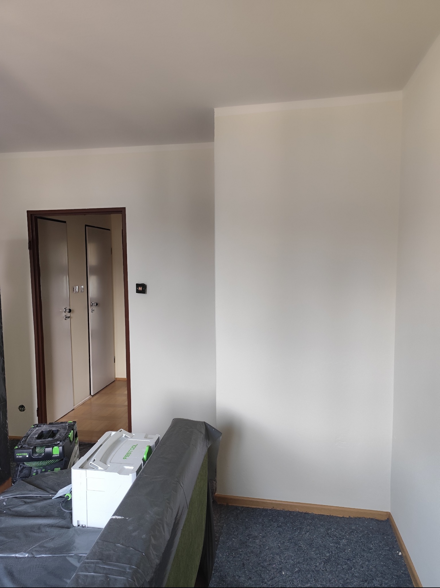 Świeżo pomalowane, jasne ściany w pokoju z widocznymi narzędziami malarskimi i zabezpieczonymi meblami. Widoczne listwy przypodłogowe i drzwi.