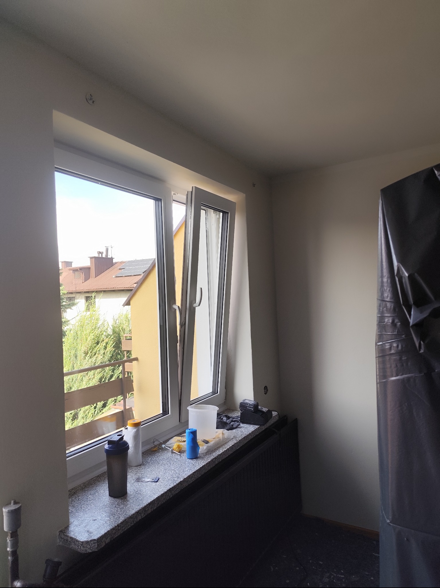 Przygotowania do malowania: okno z widokiem na zewnątrz, parapet z akcesoriami malarskimi, ściana zabezpieczona folią malarską. Widoczne ślady po gniazdkach.