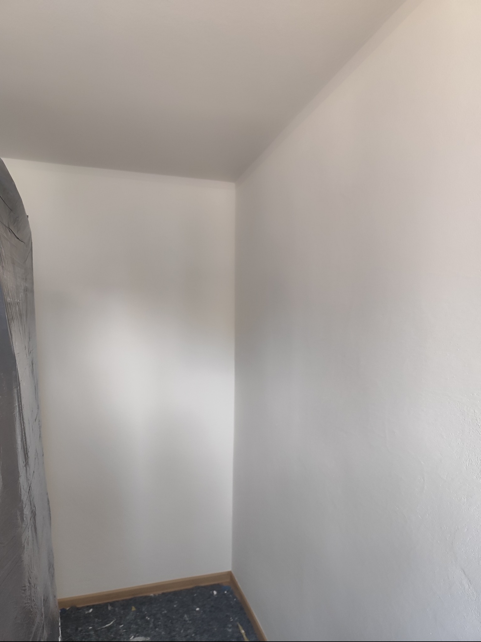 Świeżo pomalowane na biało narożne wnętrze pokoju z widocznym fragmentem zabezpieczonej folią ściany i podłogi. Listwa przypodłogowa w kolorze drewna.