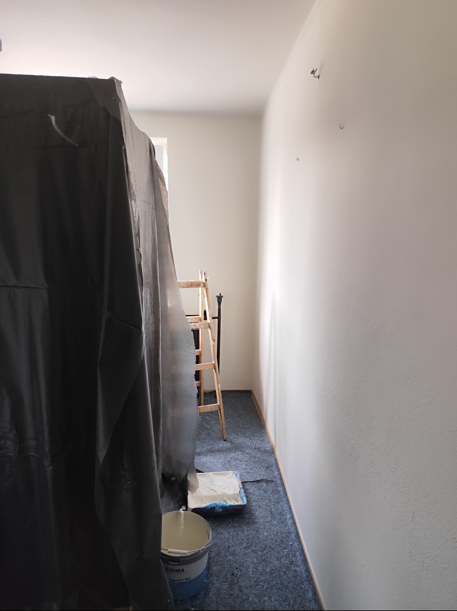 Przygotowanie do malowania: wiadro z farbą Sigma, drabina i folia ochronna w jasnym pokoju z pomalowaną ścianą. Widoczne ślady po hakach na ścianie.
