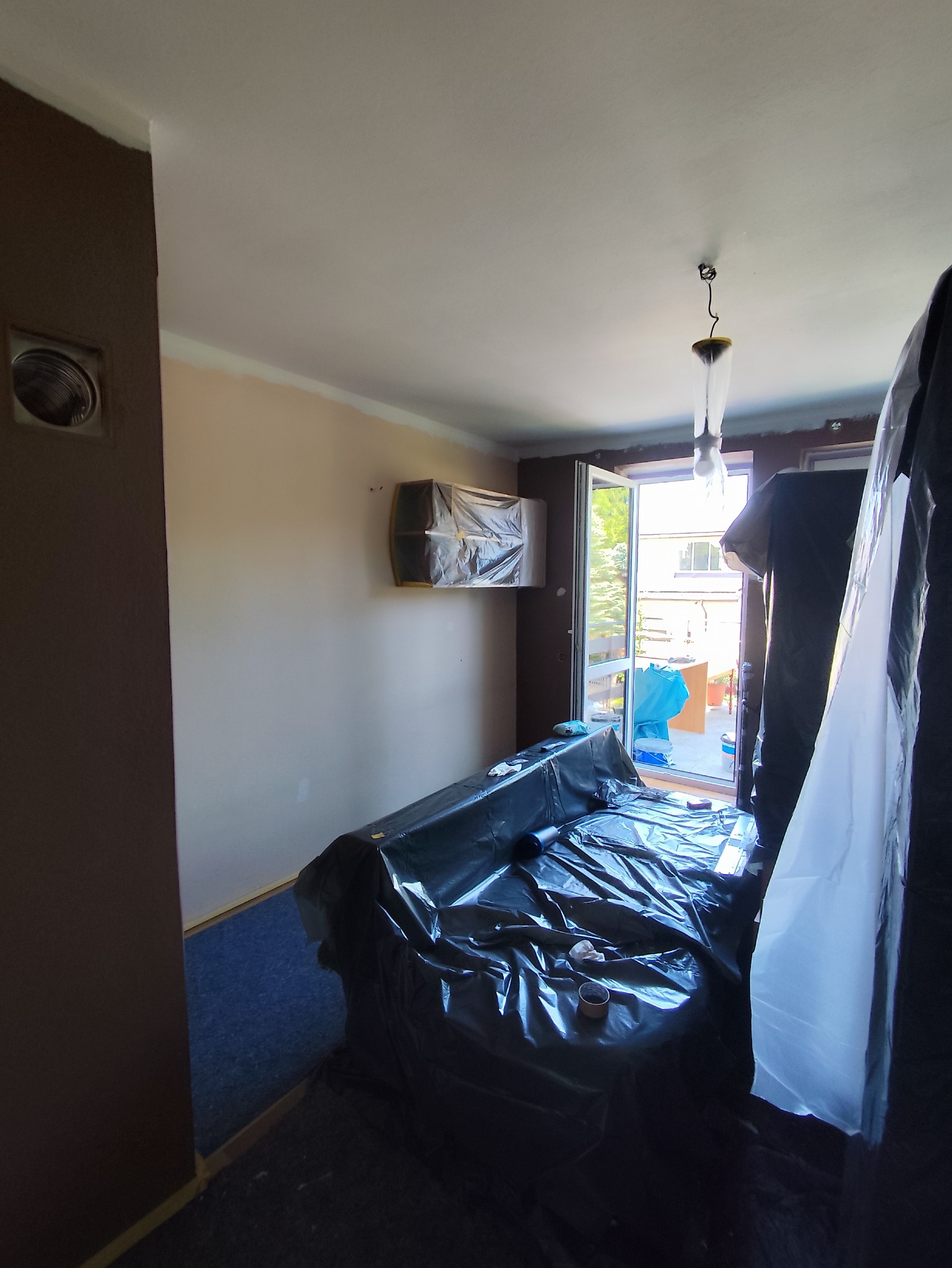 Pokój w trakcie przygotowań do malowania: meble i podłoga zabezpieczone folią, ściany częściowo pomalowane, widoczne taśmy malarskie przy suficie.