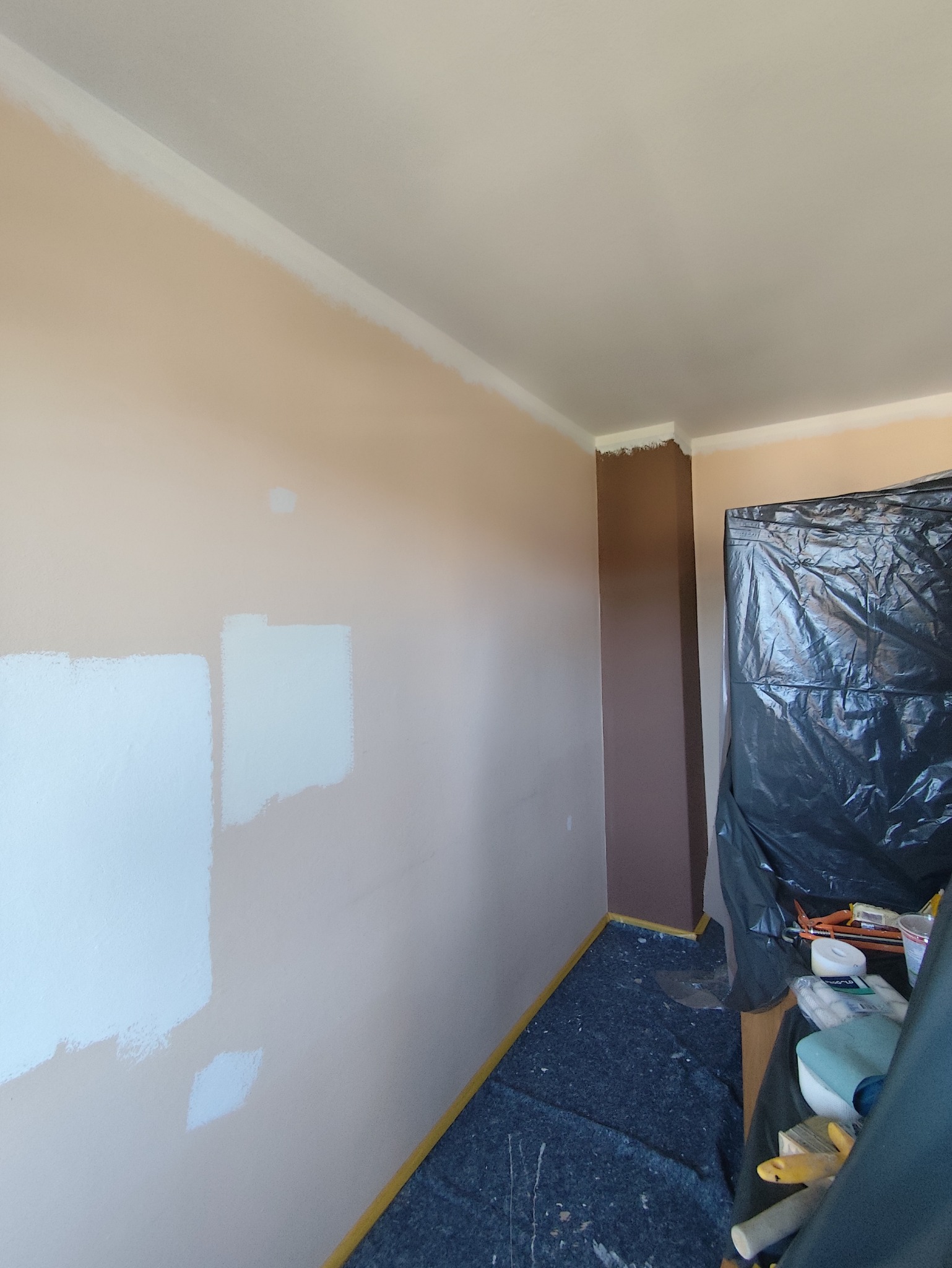 Pokój w trakcie malowania: ściany w beżowym odcieniu z fragmentami białej farby testowej, zabezpieczona podłoga, widoczne narzędzia malarskie.