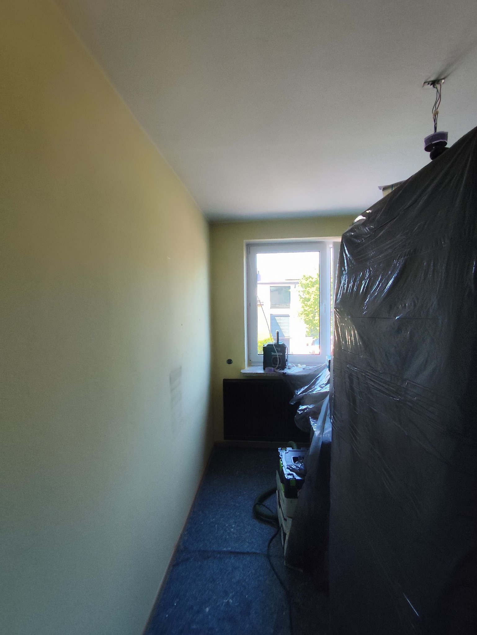 Pokój po malowaniu ścian na jasnożółty kolor. Okno z widokiem na zewnątrz. Podłoga zabezpieczona folią. Sprzęt malarski przykryty folią ochronną.