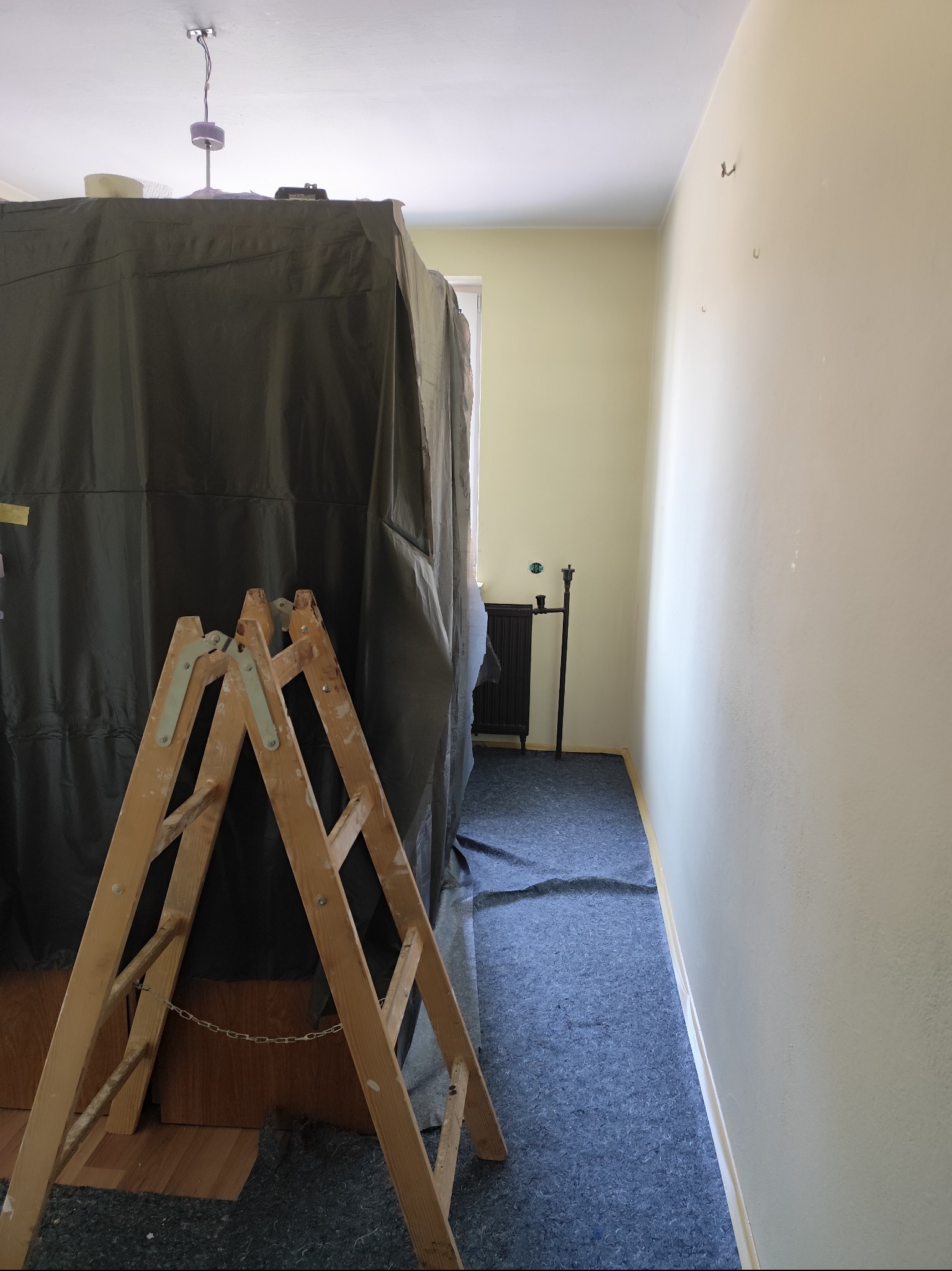 Pokój w trakcie przygotowań do malowania: meble zakryte folią ochronną, drabina malarska, zabezpieczona podłoga. Widoczne ślady po hakach na ścianie.