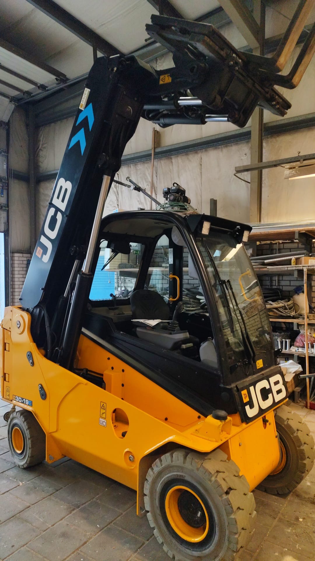 Żółty wózek widłowy JCB z czarnym masztem i chwytakiem do płyt, stojący w magazynie z widocznymi półkami i narzędziami w tle. Widok z boku, lekko od dołu.