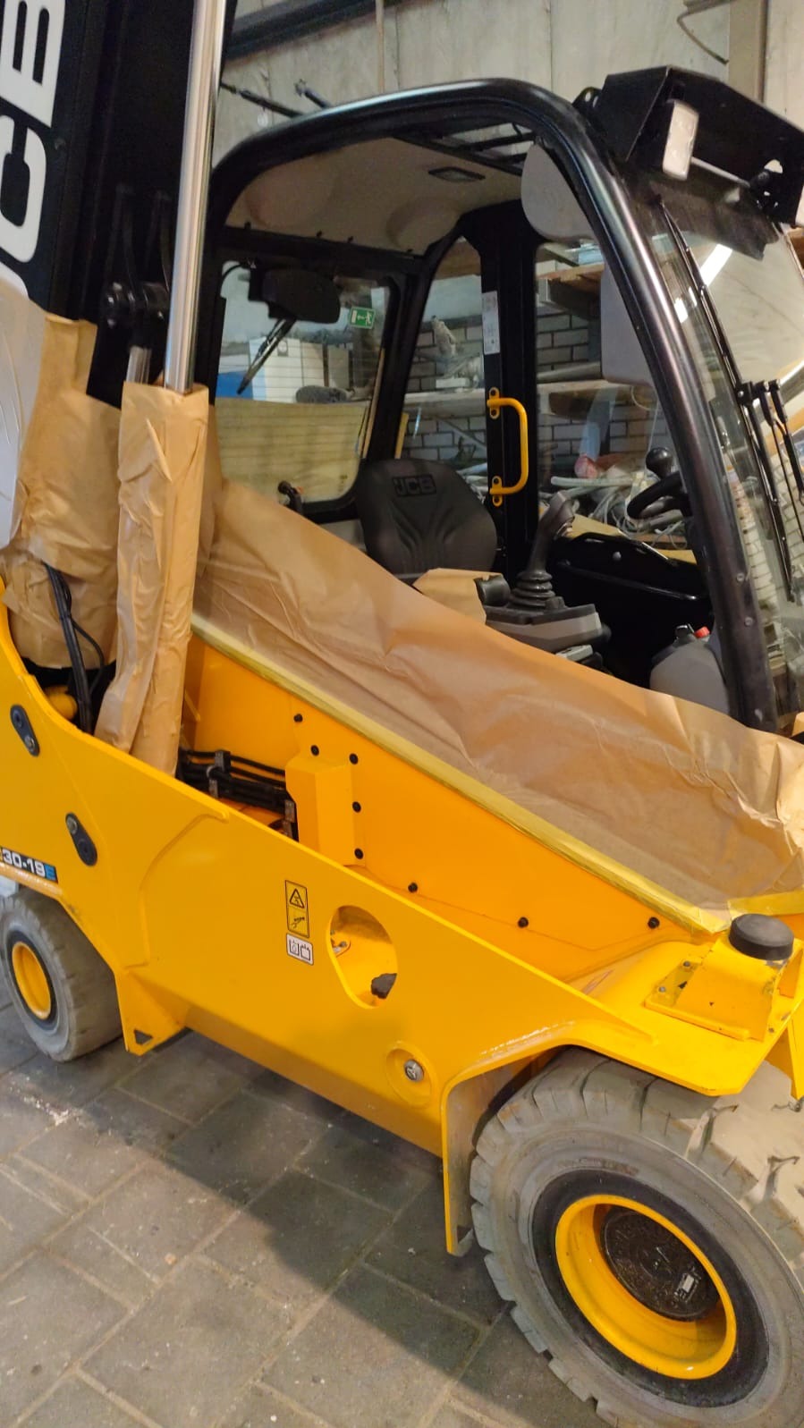 Żółty wózek widłowy JCB zabezpieczony papierem malarskim przed malowaniem. Widoczne opony, kabina operatora i elementy hydrauliczne. Przygotowanie do lakierowania.