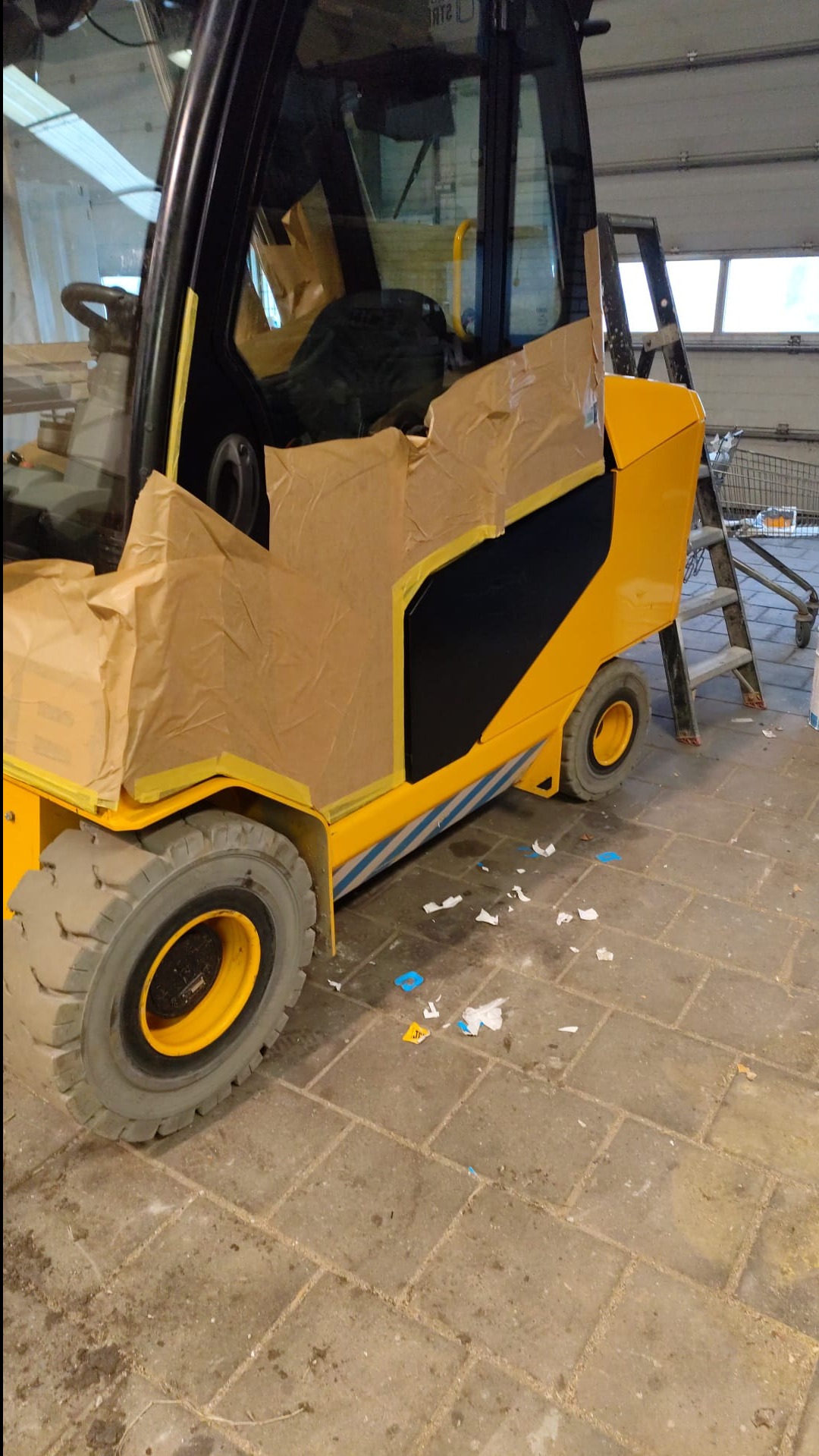 Żółty wózek widłowy, częściowo oklejony papierem maskującym przed malowaniem, stoi na betonowej posadzce w hali. Widoczna drabina i wózek sklepowy w tle.