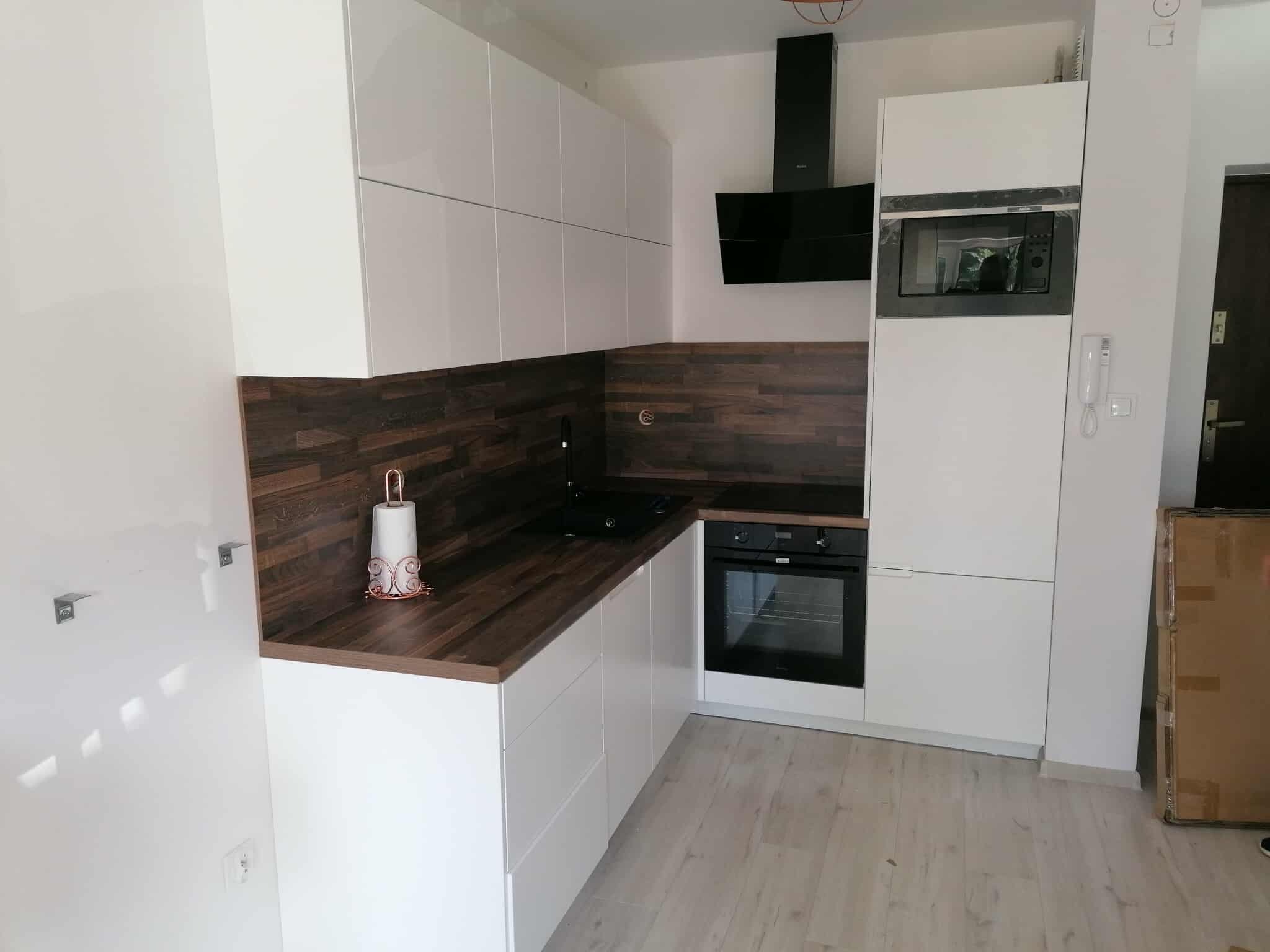 Nowoczesna kuchnia z białymi szafkami i blatem z ciemnego drewna. Czarny okap i piekarnik w zabudowie. Minimalistyczny design, jasna podłoga.