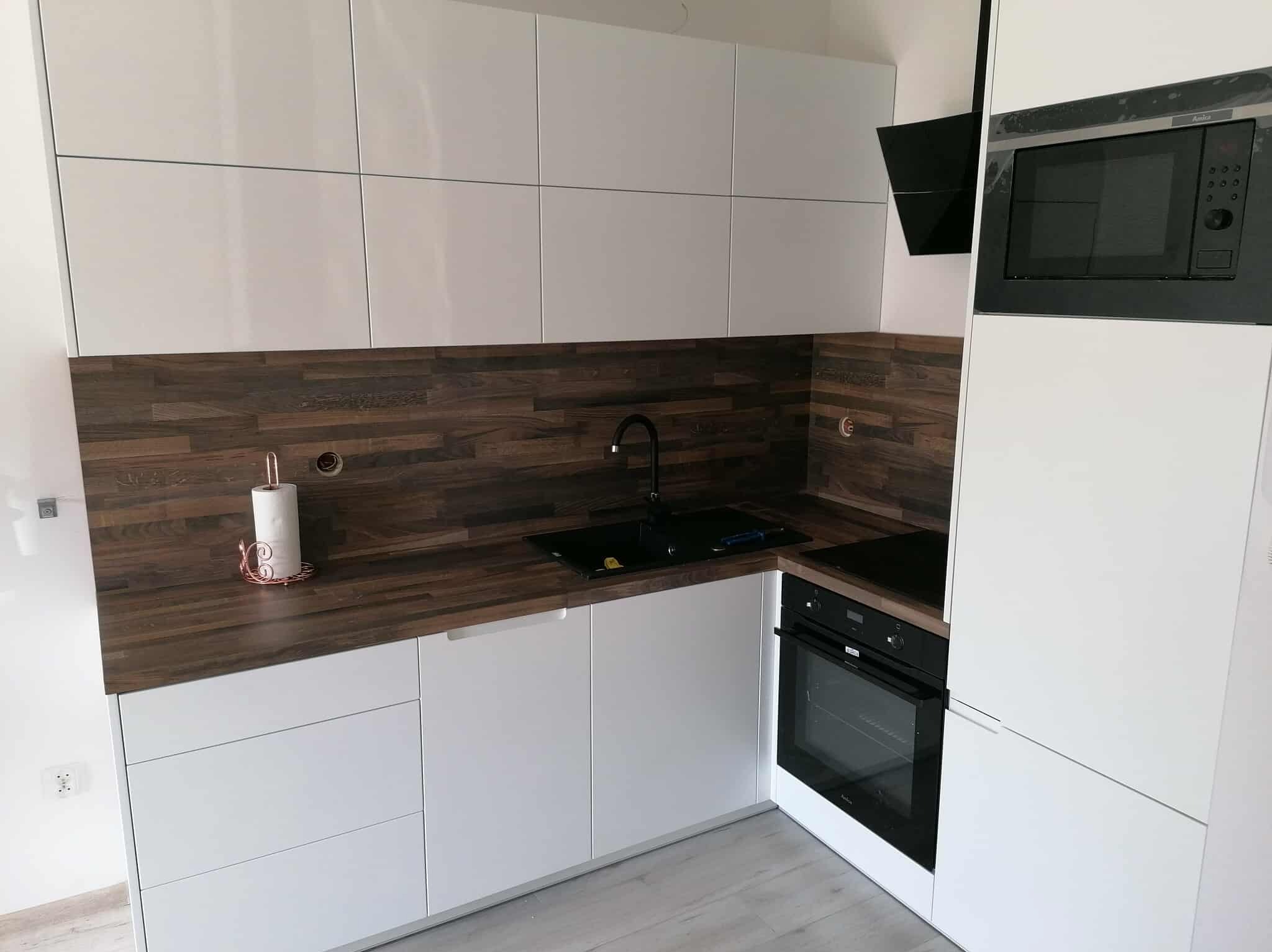 Nowoczesna kuchnia z białymi szafkami i blatem imitującym drewno. Czarny zlew i bateria, piekarnik w zabudowie. Minimalistyczny design, jasne wnętrze.