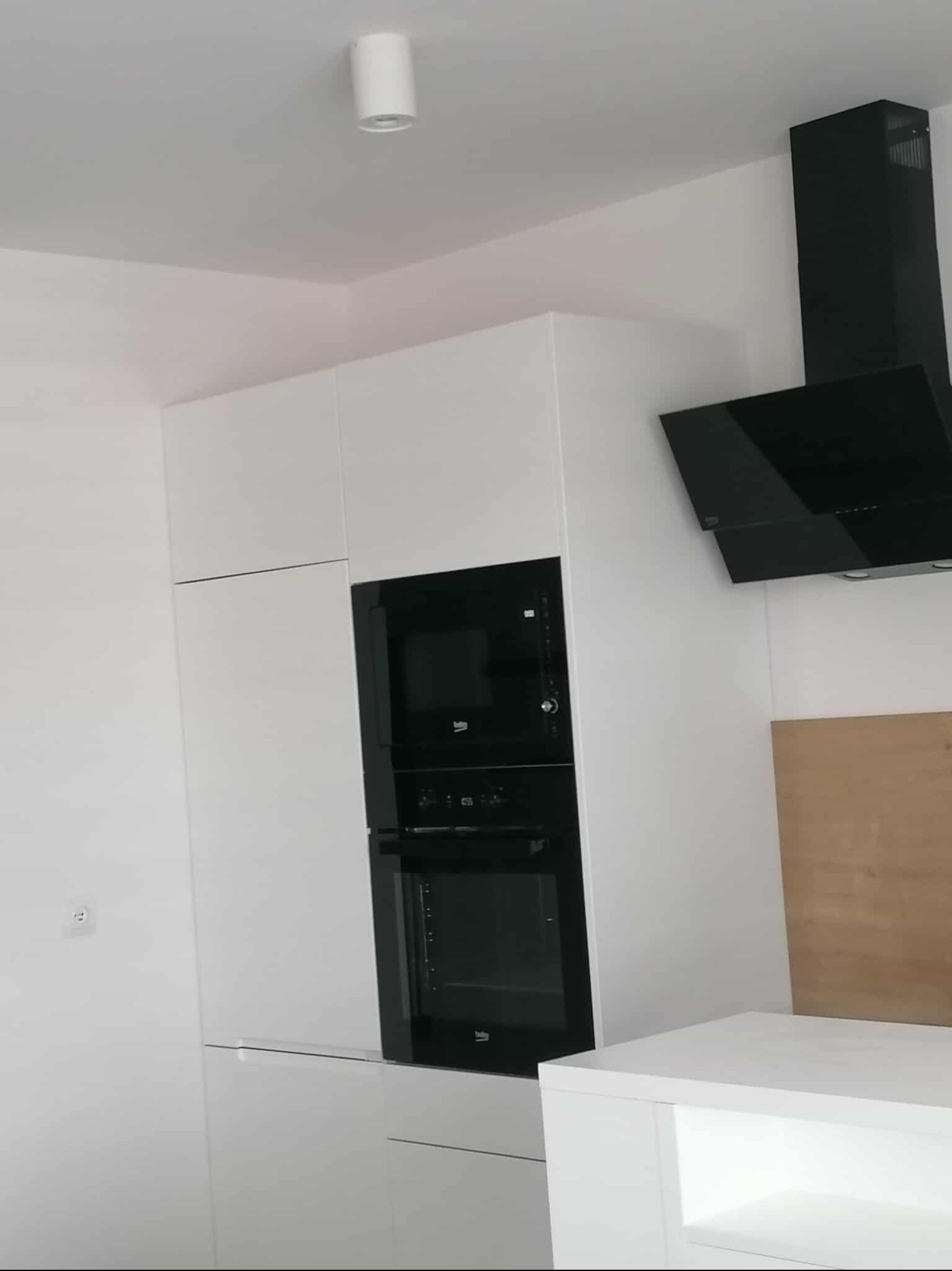 Biała zabudowa kuchenna z piekarnikiem i mikrofalówką w czarnych obudowach, uzupełniona o czarny okap. Minimalistyczny design, nowoczesne wykończenie.