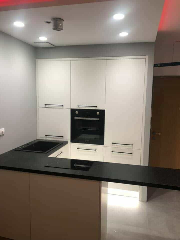 Nowoczesna kuchnia z białymi szafkami, czarnym blatem i wbudowanym piekarnikiem. Minimalistyczny design, oświetlenie sufitowe LED, czarny zlew i płyta indukcyjna.