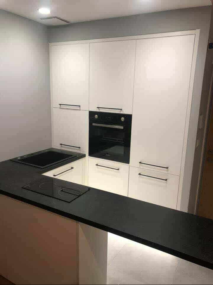 Nowoczesna kuchnia z białymi szafkami, czarnym blatem i wbudowanym piekarnikiem. Minimalistyczny design, oświetlenie LED pod blatem. Ujęcie z perspektywy.