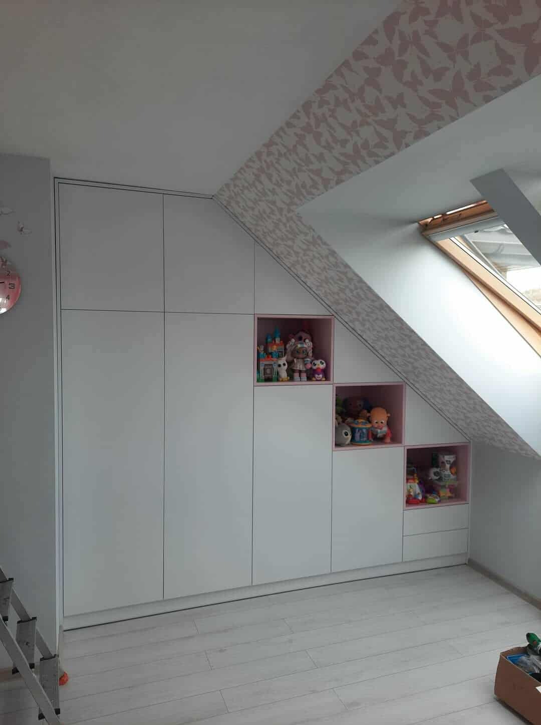 Biała zabudowa szafy wnękowej na poddaszu ze schodkowymi, różowymi wnękami na zabawki. Widoczne okno dachowe i wzorzysta tapeta w motyle.
