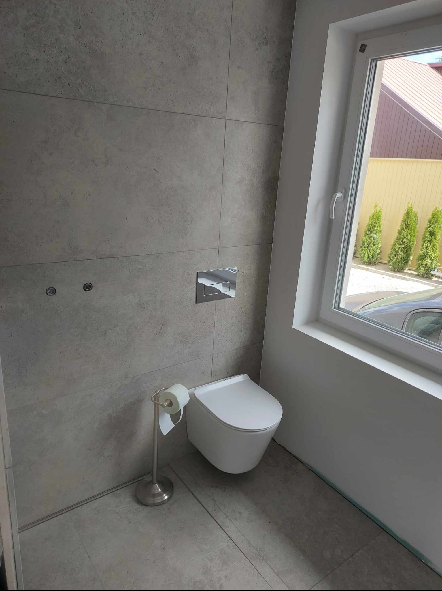 Wykończona łazienka z szarymi płytkami, wiszącą toaletą i oknem. Widoczne detale: uchwyt na papier toaletowy i przycisk spłuczki. Minimalistyczny design.