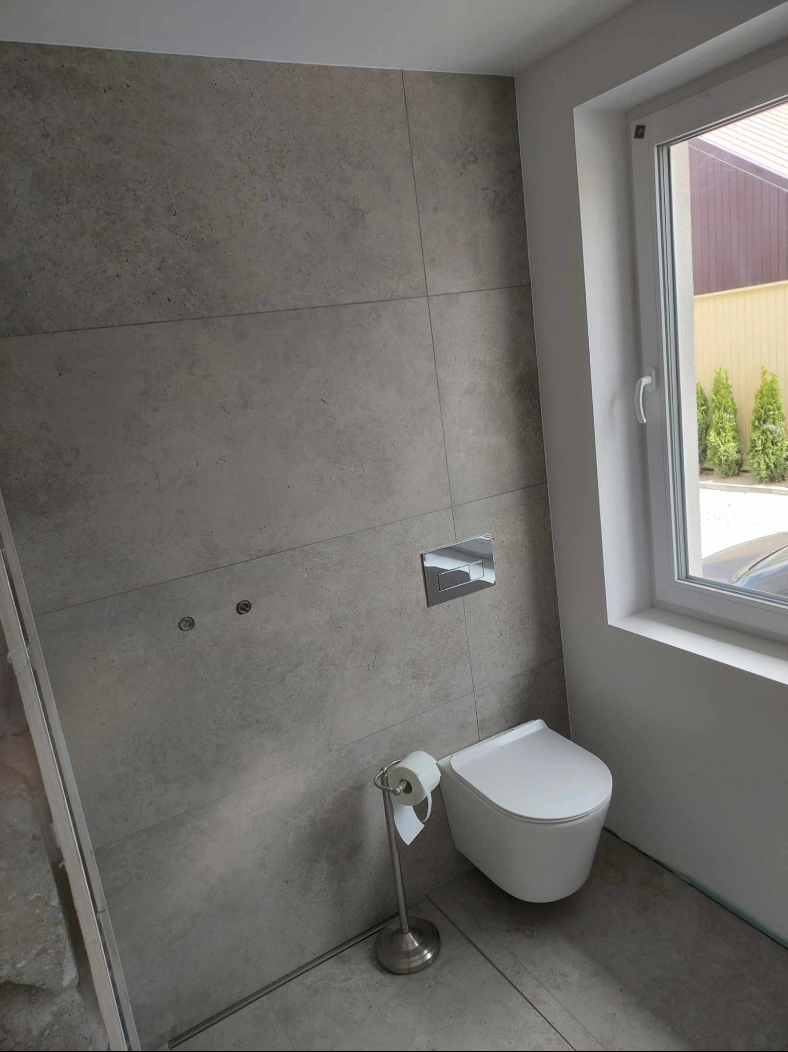 Nowoczesna toaleta z szarymi płytkami wielkoformatowymi, podwieszana miska WC i chromowany przycisk spłukujący. Widok na okno z zielonymi tujami w tle.