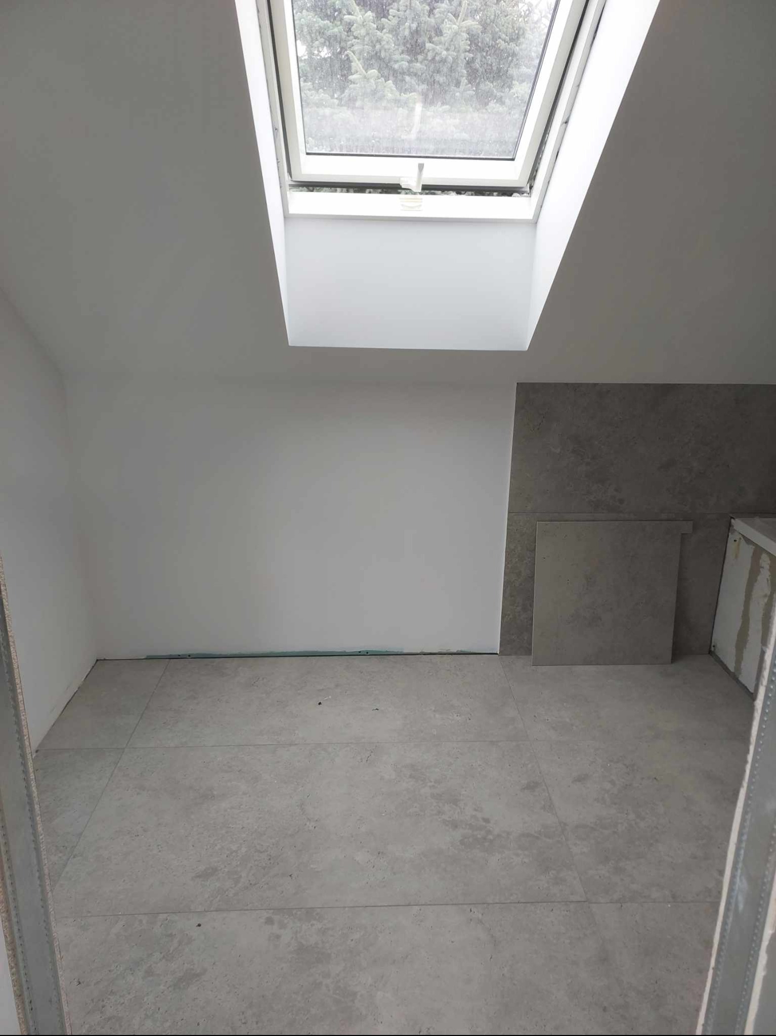 Jasne płytki podłogowe w pokoju z oknem dachowym, ściany częściowo pomalowane na biało, częściowo wyłożone szarymi płytkami. Widoczne krawędzie płyt i niedokończone detale.