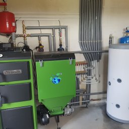 HYDRAULIK Meik Barwinski - Zestaw kotłów: czarny Kamen i zielony z logo, obok biały zbiornik Keller. Szare rury instalacji grzewczej na ścianie. Całość w pomieszczeniu kotłowni.