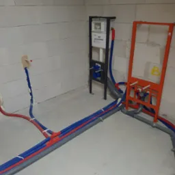 Instalacja wodno-kanalizacyjna w surowym stanie: rury w izolacji czerwono-niebieskiej, stelaż podtynkowy WC, szara kanalizacja, ściany z betonu komórkowego.