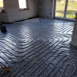 HYDRAULIK Meik Barwinski - Ułożone rury ogrzewania podłogowego na folii z nadrukowaną siatką w surowym wnętrzu. Widoczne okna i narzędzia.