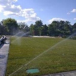 HYDRAULIK Meik Barwinski - Działający system nawadniania trawnika z zieloną skrzynką sterującą, widok z boku na osobę siedzącą na obrzeżu, w tle drzewa i błękitne niebo z chmurami.