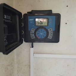 HYDRAULIK Meik Barwinski - Sterownik nawadniania Orbit z otwartą obudową, zamontowany na ścianie. Widoczny panel sterowania z wyświetlaczem ustawionym na godzinę 5:00 PM. Kabel w listwie przypodłogowej.