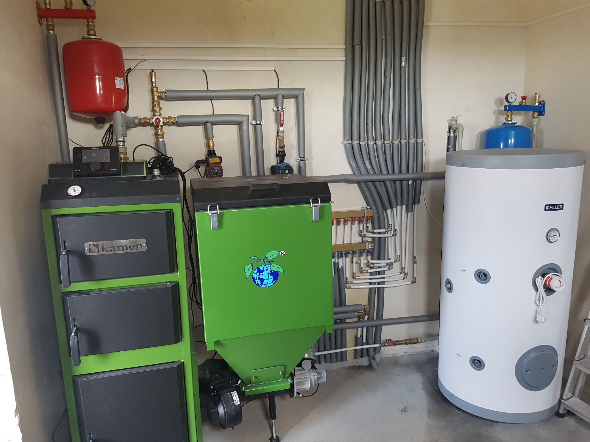 Zestaw kotłów: czarny Kamen i zielony z logo, obok biały zbiornik Keller. Szare rury instalacji grzewczej na ścianie. Całość w pomieszczeniu kotłowni.