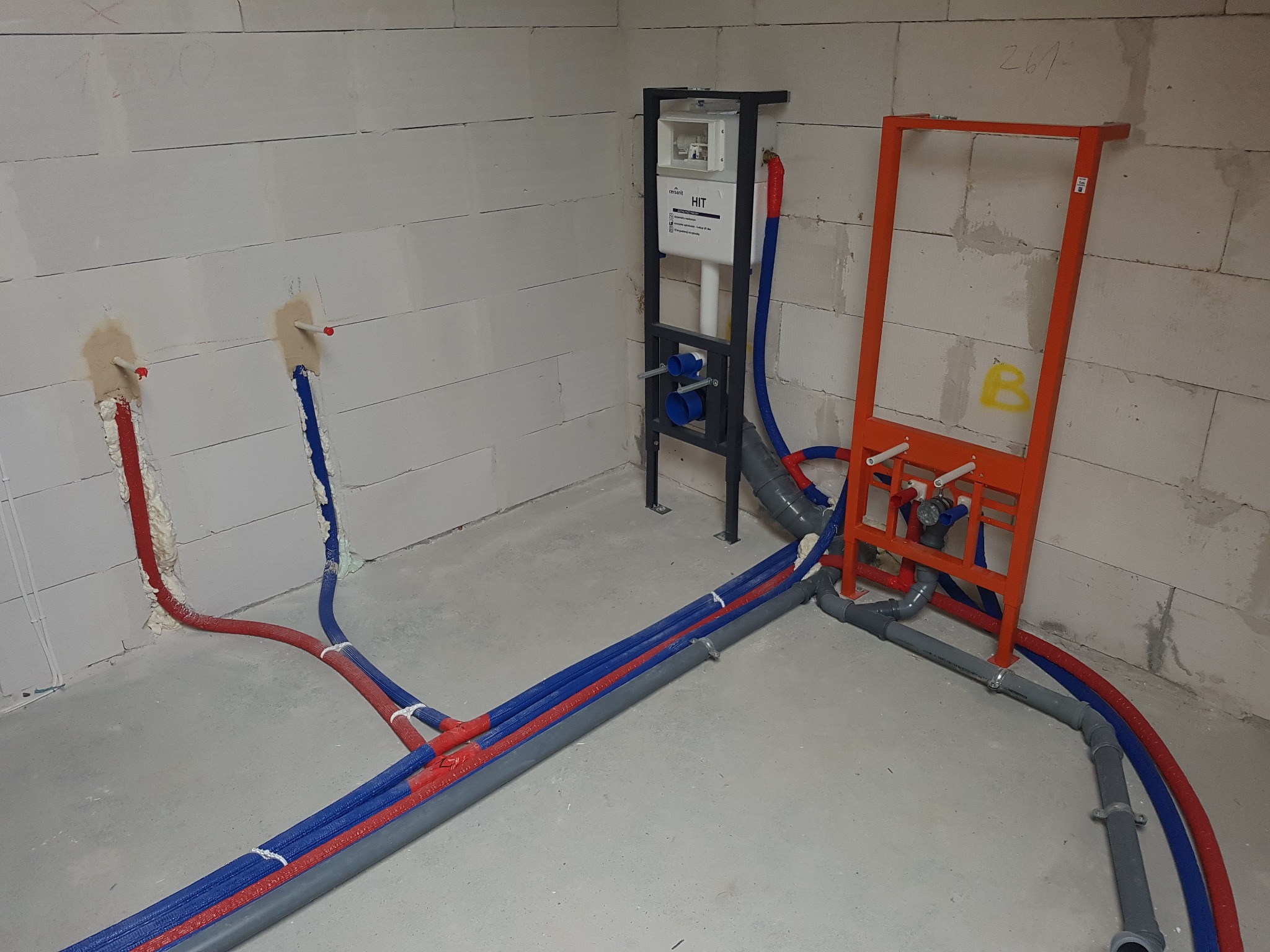 Instalacja wodno-kanalizacyjna w surowym stanie: rury w izolacji czerwono-niebieskiej, stelaż podtynkowy WC, szara kanalizacja, ściany z betonu komórkowego.