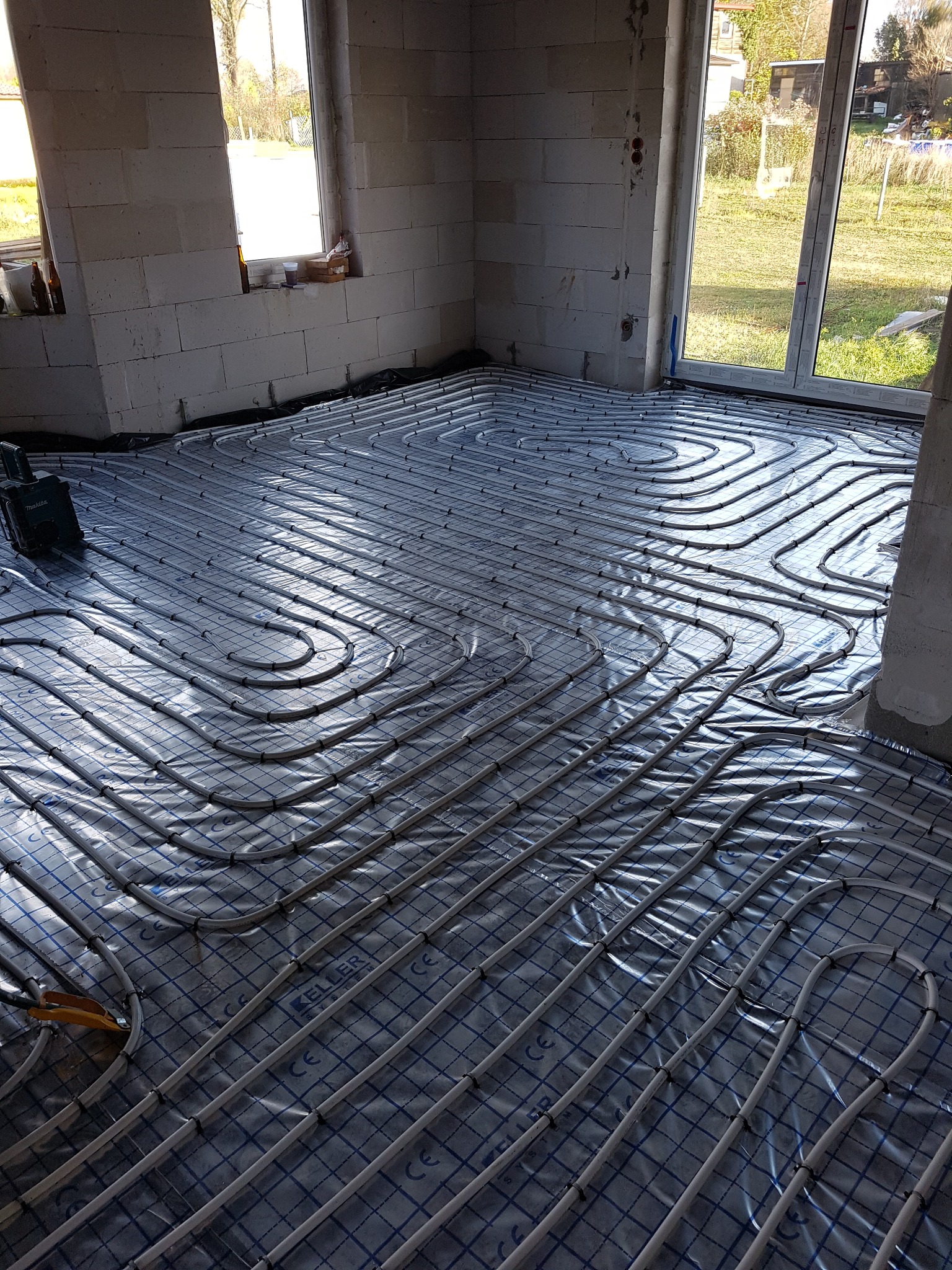 Ułożone rury ogrzewania podłogowego na folii z nadrukowaną siatką w surowym wnętrzu. Widoczne okna i narzędzia.