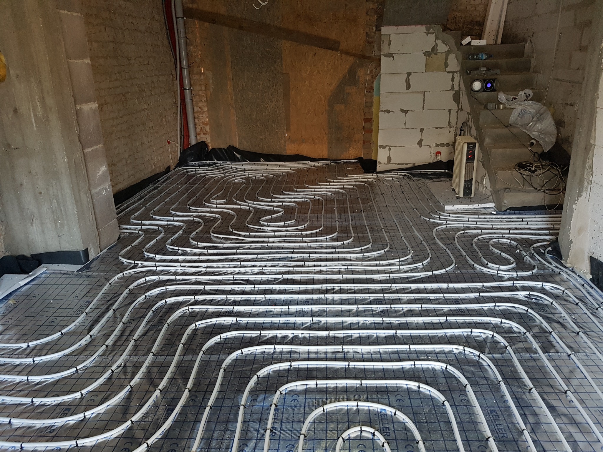 Instalacja ogrzewania podłogowego z białych rur na folii z siatką, widok z góry. W tle surowe ściany i schody w trakcie budowy.
