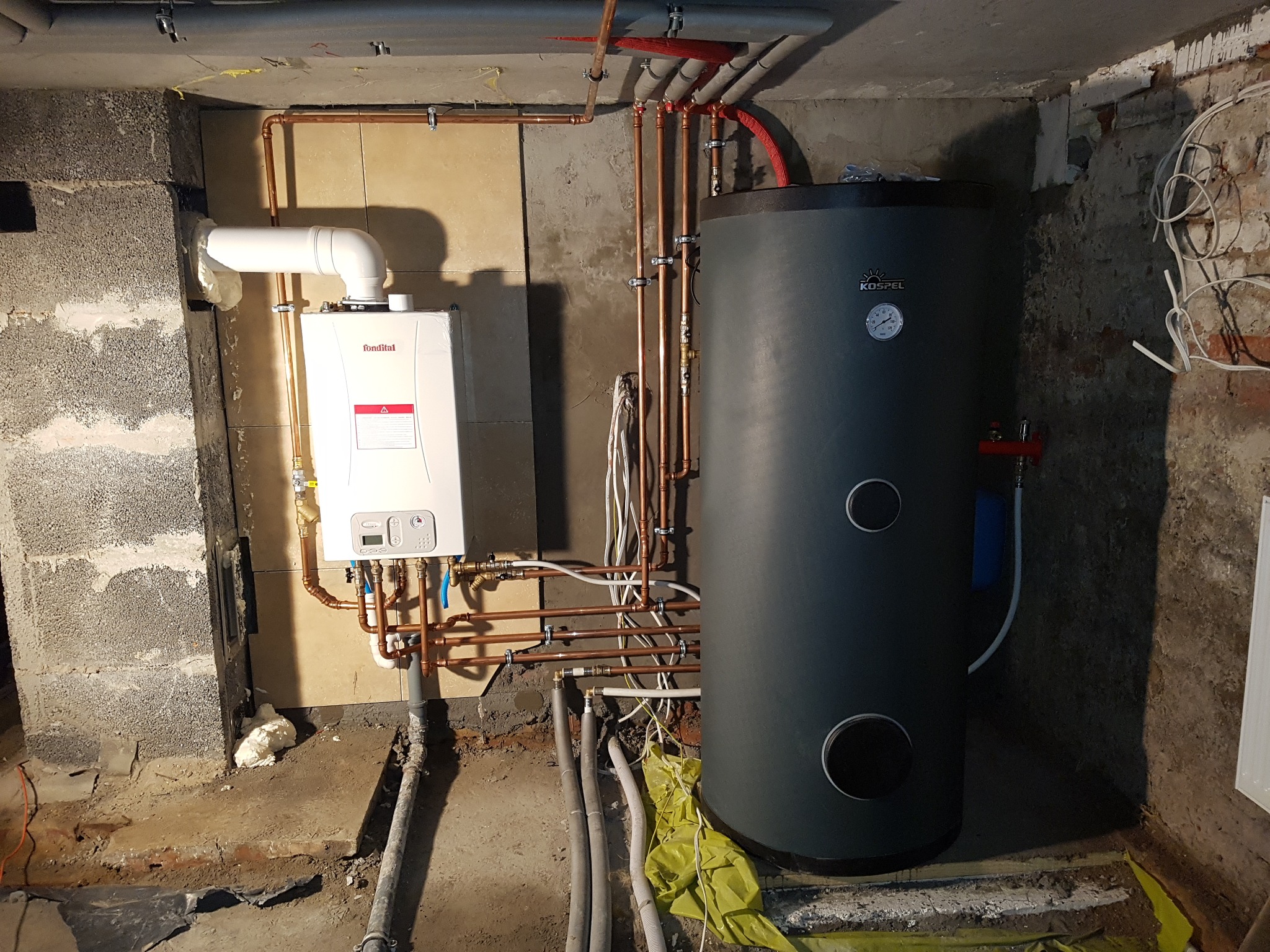Zainstalowany gazowy piec dwufunkcyjny Fondital obok szarego zasobnika Kospel, połączone miedzianymi rurami w surowym pomieszczeniu z odsłoniętymi instalacjami.