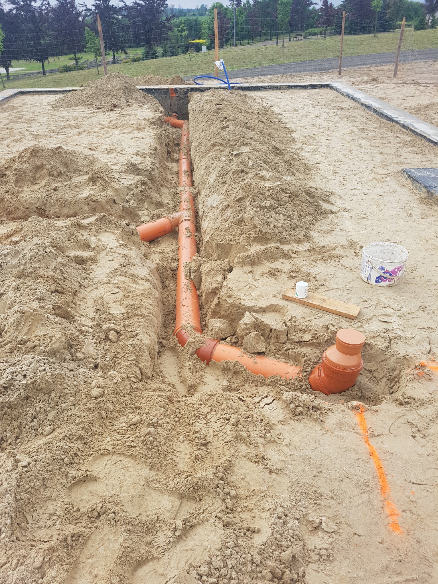 Pomarańczowe rury kanalizacyjne ułożone w wykopie wypełnionym piaskiem. Widoczne złączki i kształtki, etap prac ziemnych. W tle trawa i drzewa.
