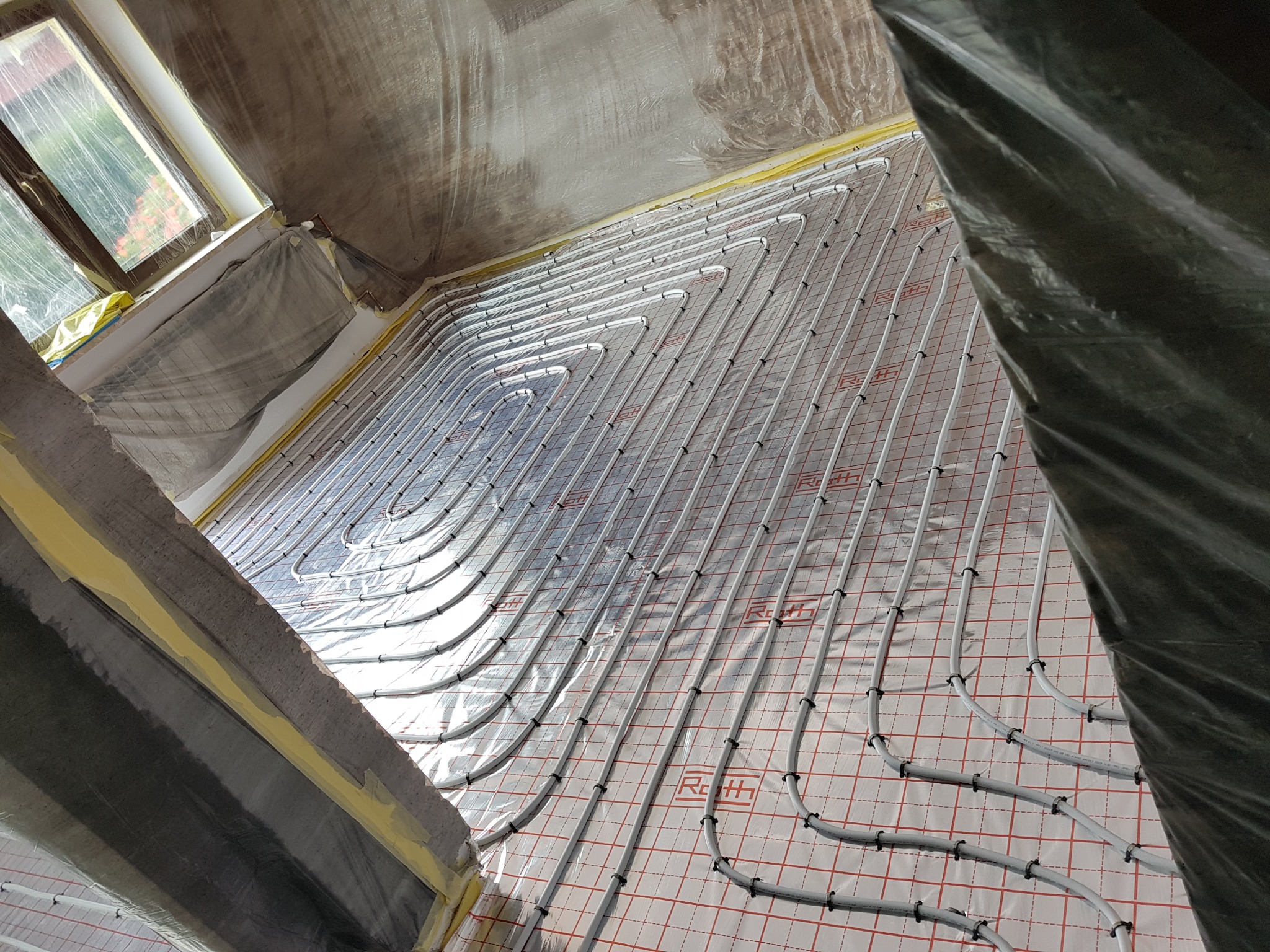 Ułożone rury ogrzewania podłogowego marki Roth na folii z czerwoną kratką, widoczne mocowania, okno z folią ochronną, przygotowane do zalania posadzką.
