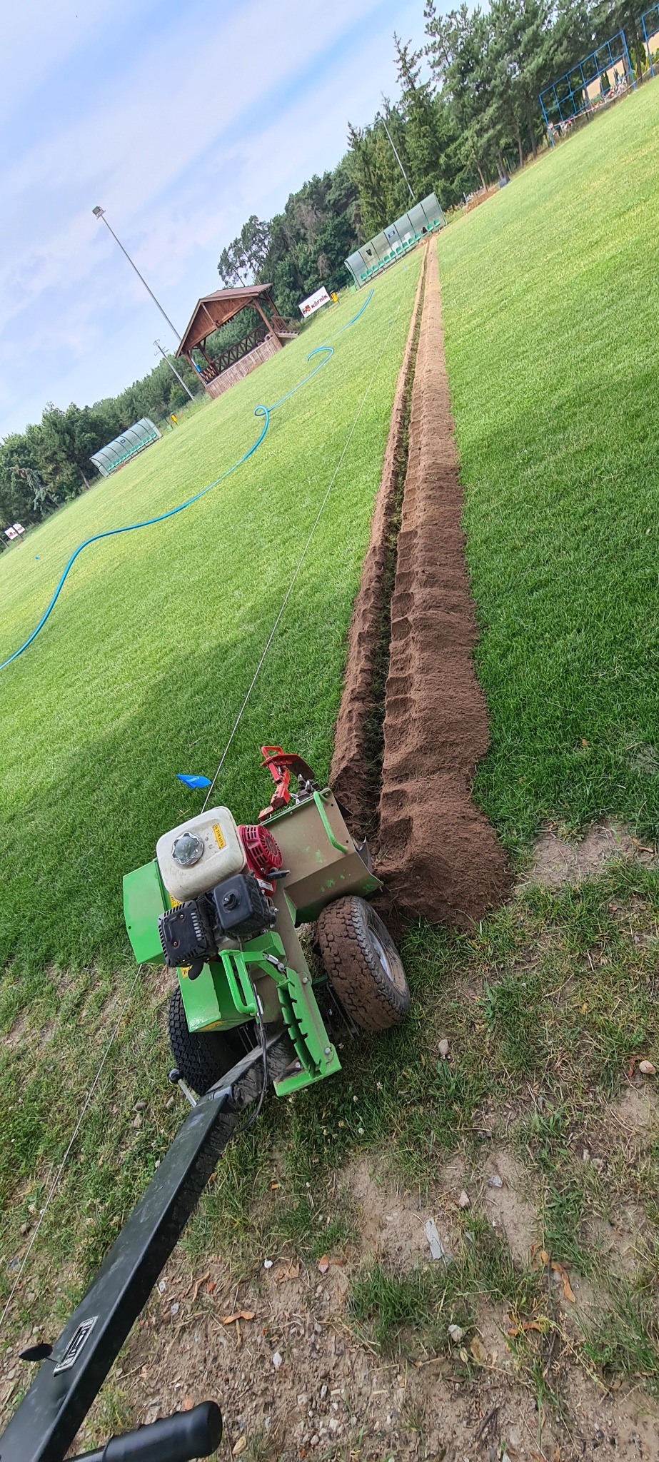 Wykonanie rowu pod system nawadniania na trawniku za pomocą specjalistycznej maszyny. W tle widoczna altana i drzewa.