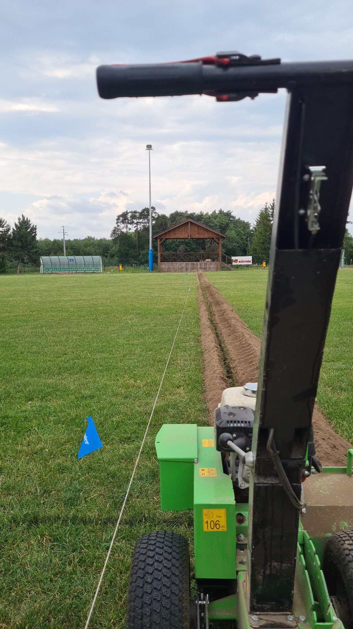 Zielona maszyna wykopuje wąski rów w trawie, wyznaczony sznurkiem i niebieskim znacznikiem, prawdopodobnie pod system nawadniania boiska sportowego.