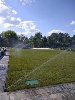 Działający system nawadniania trawnika z zieloną skrzynką sterującą, widok z boku na osobę siedzącą na obrzeżu, w tle drzewa i błękitne niebo z chmurami.