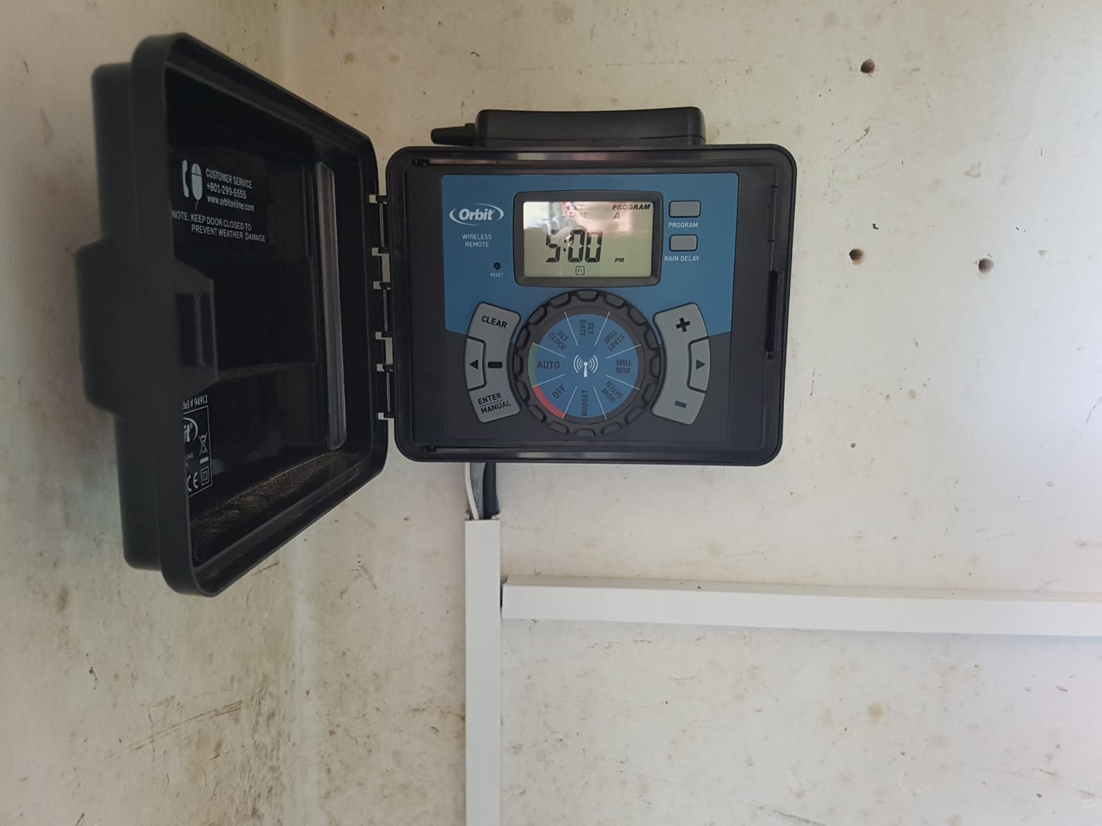 Sterownik nawadniania Orbit z otwartą obudową, zamontowany na ścianie. Widoczny panel sterowania z wyświetlaczem ustawionym na godzinę 5:00 PM. Kabel w listwie przypodłogowej.