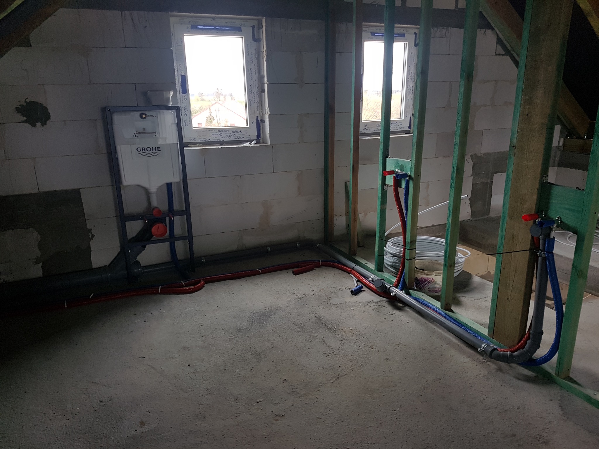 Instalacja wodno-kanalizacyjna w surowym wnętrzu. Widoczne rury, stelaż podtynkowy Grohe, okna i surowe ściany z betonu komórkowego. Prace instalacyjne w toku.