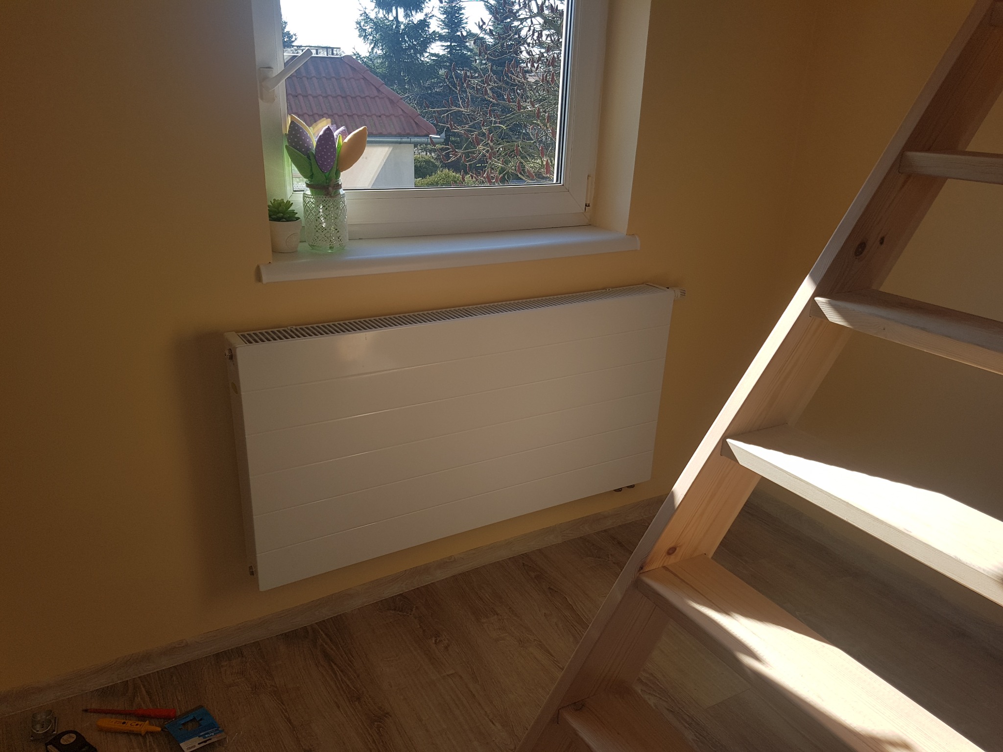 Nowy, biały grzejnik panelowy pod oknem w pokoju z drewnianą drabiną, narzędzia na podłodze. Widok na zewnątrz przez okno. Słoneczny dzień.